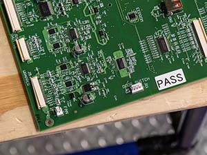 Electronic circuit board with a 'PASS' inspection label laying on a wood table.
