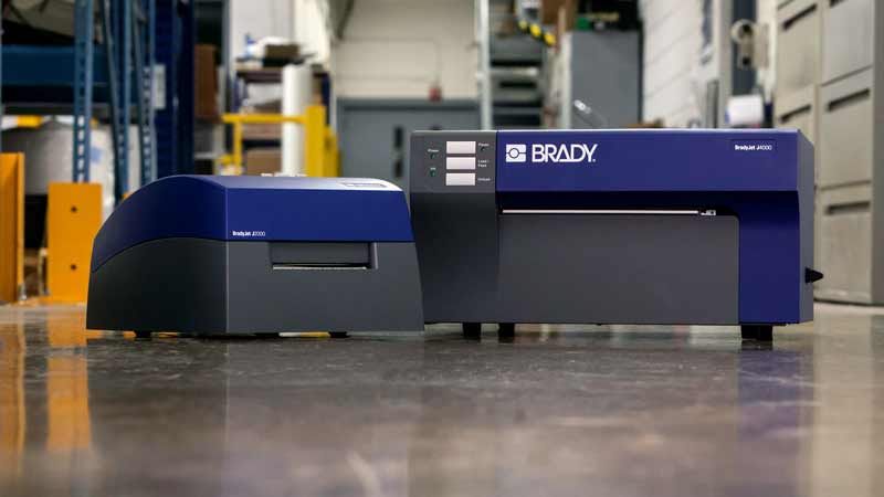 A Brady J3000 and J4000 Inkjet benchtop printer sitting on a table in a warehouse setting.