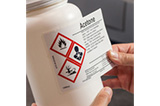 A person applying a GHS label to an acetone container.